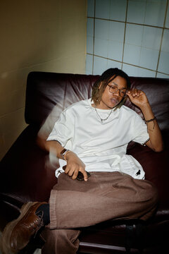 Vertical shot of young Black man sitting on sofa, holding smartphone and touching glasses, expressing fatigue and contemplation. Suitable for mental health, stress, burnout, lifestyle campaigns
