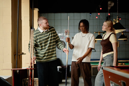 Young adult friends playing billiards, discussing cue technique during social game in pool hall. Useful for leisure marketing, youth lifestyle campaigns, teamwork concepts
