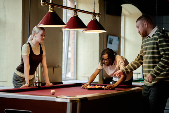 Young adult friends gathering around pool table, arranging balls and preparing game in social leisure venue. Image supporting teamwork, friendship, youth lifestyle, recreation marketing

