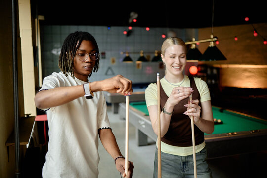 Young adult Black man and Caucasian woman preparing pool cues in billiards club, sharing leisure activity and teamwork. Useful for friendship, social lifestyle, recreation marketing
