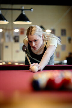 Vertical shot of Caucasian young woman aiming cue stick over pool table, focusing on precision and leisure competition. Suitable for recreation marketing, nightlife promotion, social lifestyle content