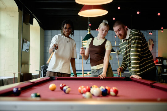 Multiethnic young adults playing billiards and aiming shots together in social club, showing teamwork and leisure bonding. Useful for lifestyle marketing, friendship, youth culture campaigns
