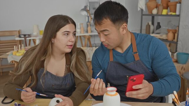 Man showing smartphone to woman who holds paintbrush and ceramic vase, both wearing aprons at a pottery studio with shelves of pots and brushes; collaboration focused.