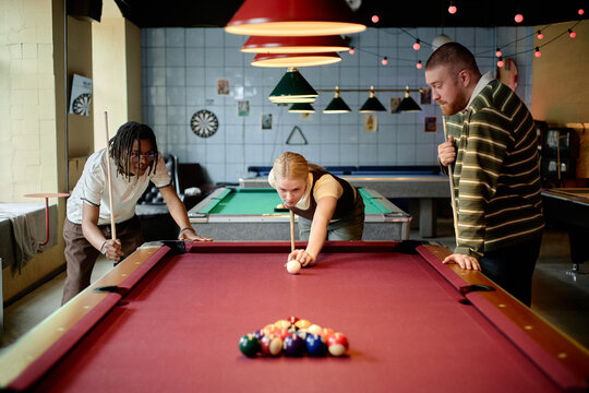 Young adult diverse friends playing billiards in recreation club, aiming cue ball and watching turn. Image supporting leisure marketing, social lifestyle campaigns, team bonding promotion

