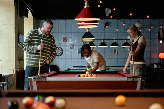 Multiethnic young adults playing billiards in pool hall, socializing during leisure time and aiming cue shot. Useful for friendship, nightlife, team bonding, recreation marketing
