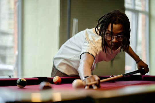 Young Black man aiming pool shot on billiards table, practicing leisure game with focus and precision. Suitable for recreation marketing, youth lifestyle, social venue promotion
