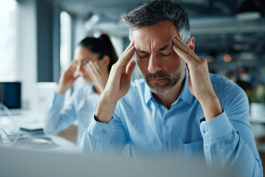 Tired man pressing temples with headache while sitting at desk, colleague offering support nearby, modern office interior softly blurred, natural daylight illumination,
