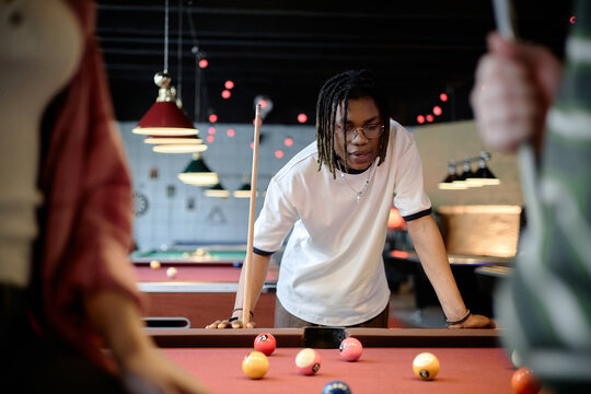 Young Black man planning pool shot with friends in recreation venue, showing teamwork and leisure socializing. Useful for youth lifestyle, friendship, entertainment marketing campaigns
