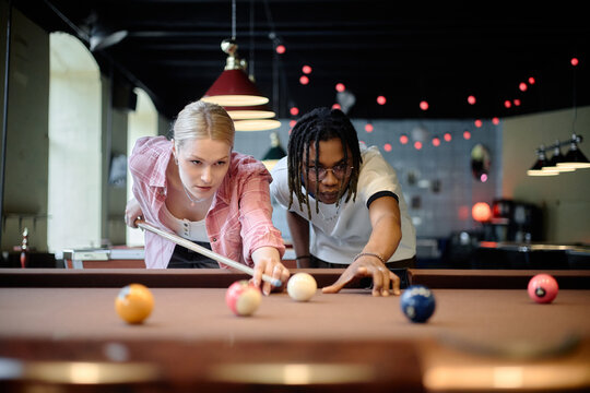 Young adult Caucasian woman and Black man playing billiards, aiming pool shot together in social venue. Supporting leisure marketing, friendship, dating, youth lifestyle campaigns
