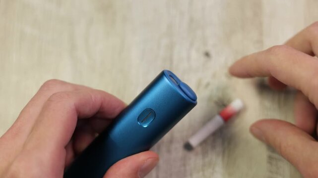 Man hand inserting a nicotine tobacco stick into a blue electronic heating device and pressing the button to start the heating process for smoking.