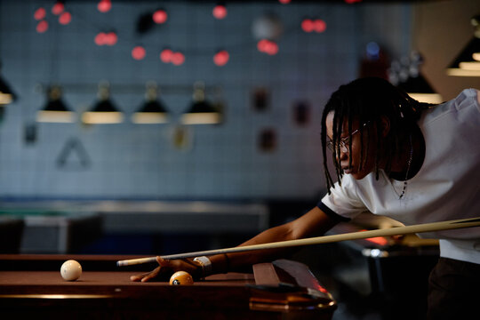 Black young adult man aiming pool cue over billiards table, focusing on precision shot during leisure game. Ideal for recreation marketing, nightlife promotion, competitive hobby content
