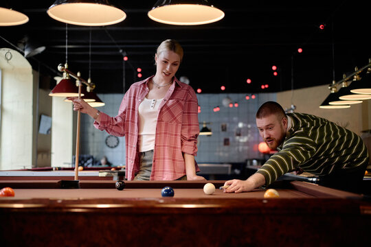 Young adult Caucasian couple playing billiards in leisure venue, man aiming shot while woman watching and holding cue. Ideal for dating, friendship, recreation, nightlife marketing
