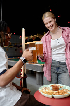 Vertical shot of young adult women clinking beer mugs while socializing beside pool table, representing friendship and leisure. Useful for nightlife marketing, bar promotion, vertical
