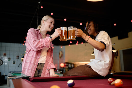 Young adult Caucasian woman and Black man clinking beer mugs while socializing over pool table, representing friendship and leisure marketing. Useful for nightlife, bar promotion, social campaign
