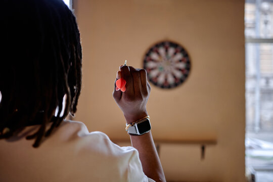 Black young adult woman aiming dart at board, practicing focus and leisure game indoors. Useful for recreation marketing, concentration training, lifestyle wellness campaigns
