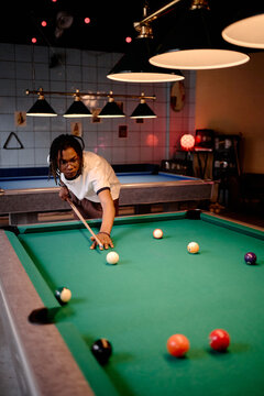 Vertical shot of young Black man aiming pool cue over billiards table, practicing precision and leisure activity. Suitable for nightlife marketing, recreation ads, social lifestyle content
