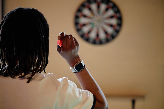 Black teenager aiming dart at board, practicing focus and hand eye coordination during indoor game. Useful for youth recreation, concentration, leisure activity, medium shot
