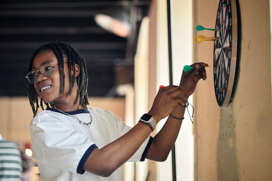 Young African American man placing dart on board, playing target game and practicing focus and precision. Useful for teamwork, leisure marketing, goals, strategy, medium shot
