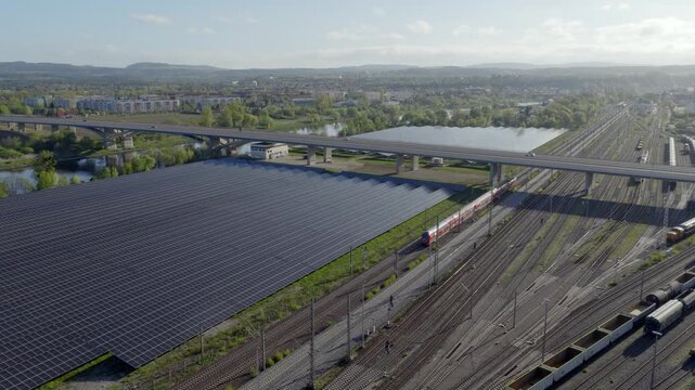 Aerial view of a large solar farm next to railway tracks with driving trains and a highway bridge, symbolizing energy transition and modern infrastructure.