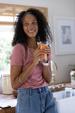 African American woman holding glass of orange juice leaning on wooden countertop in kitchen
