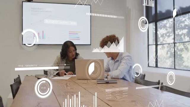 Woman writing, glancing at laptop, peer leaning, pointing, overlays moving on charts for biz review