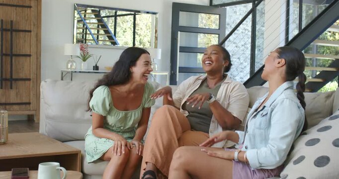 Diverse female friends laughing, reacting on couch as right woman showing story, smartwatch visible