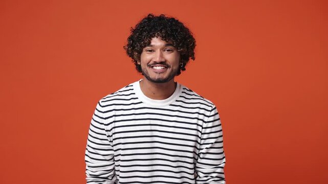 Excited cheerful young Indian man wear striped sweatshirt casual clothes showing thumbs up gesture like sign looking camera isolated on plain orange red background studio portrait. Lifestyle concept