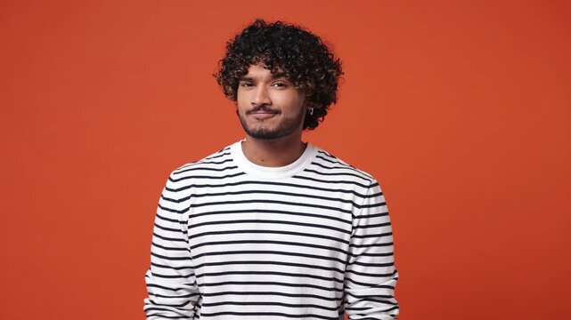 Young Indian man wear striped shirt casual clothes look aside say do not talk hush be quiet with finger on lips shhh gesture isolated on plain orange red background studio portrait. Lifestyle concept