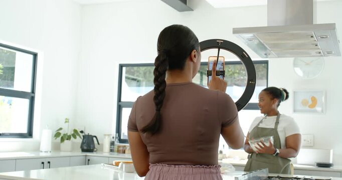 Diverse women filming recipe with ring light in kitchen, host tapping phone while cohosts mixing