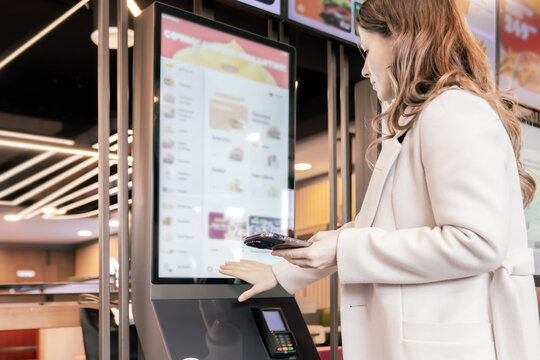 Woman using self service kiosk with smartphone for contactless food ordering and payment in modern fast food restaurant interior