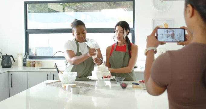 Phone filming, Diverse female bakers in aprons piping and placing fruit on two-tier cake at counter