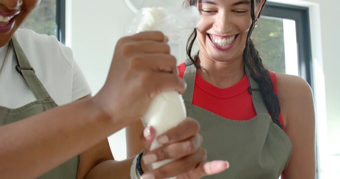 Diverse female friends in aprons squeezing bag, finishing 2-tier cake with blueberries at counter