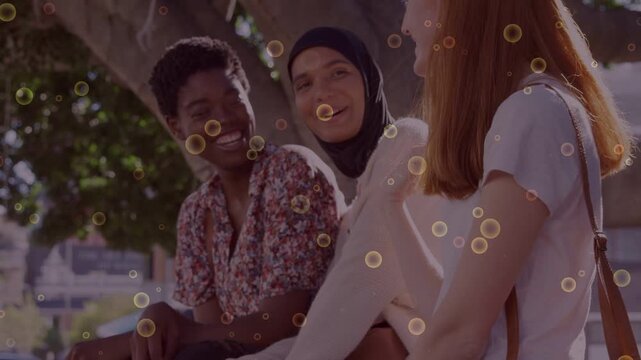 Right woman speaking, three women laughing, leaning, smiling, bubbles floating over bench for ads