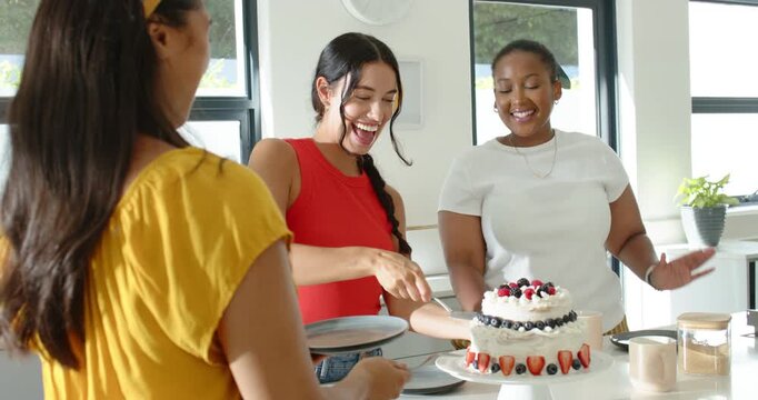 Female friends celebrating as woman in red cutting and serving cake with server on kitchen counter