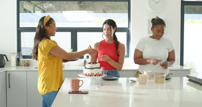 Diverse women left woman offering server, center woman cutting two tier cake at home celebrating