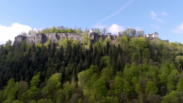 Aerial view of K&ouml;nigstein Fortress in Saxon Switzerland atop a steep sandstone rock, surrounded by dense spring forest under a blue sky.