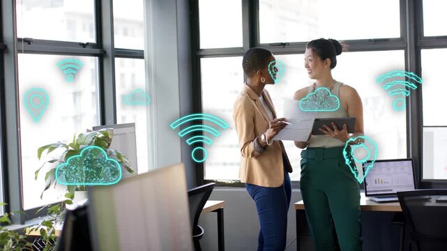 Two coworkers reviewing docs, tapping tablet, initiating cloud Wi-Fi icons over office laptop chart