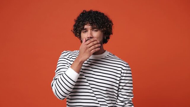 Tired sad young Indian man wear striped sweatshirt casual clothes did not get enough sleep last night after party and barely got up in the morning yawn rub eyes isolated on plain orange red background