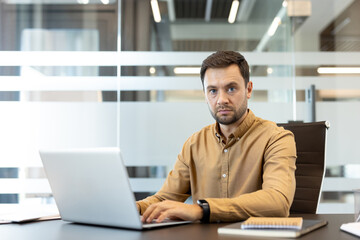 Fototapeta premium Man focusing on his laptop in a modern corporate office, looking directly at the camera with a serious expression while engaged in daily professional tasks and online communication