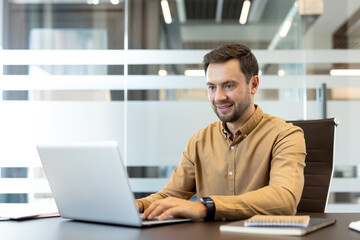 Fototapeta premium Young businessman typing on a laptop, working at a modern office desk and smiling confidently, representing productivity, success, and professional growth in a corporate environment