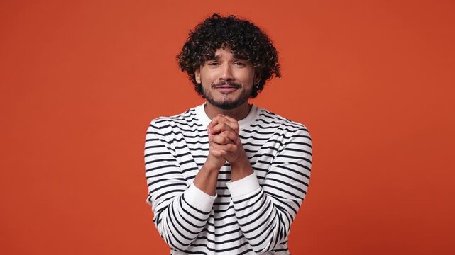 Pleading worried young Indian man wear striped sweatshirt casual clothes hold hands folded in prayer begging about something making wish keep fingers crossed isolated on plain orange red background