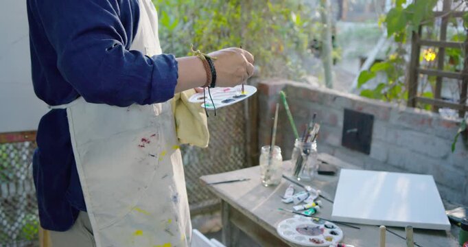 Senior Asian man apron holding palette, brush, mixing paint for blank canvas on terrace, copy space