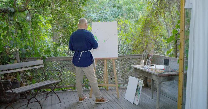 Senior Asian man in apron at easel on deck, starting blank canvas, painting tree-form with brush