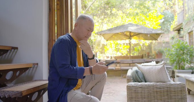Senior Asian man sitting on wood bench checking phone, tapping, scrolling, glancing toward garden