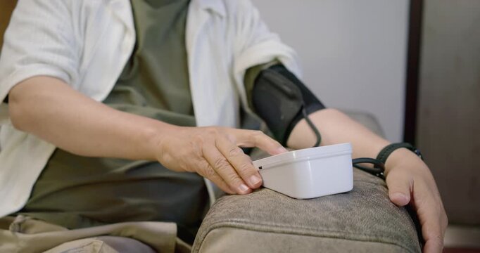 Senior Asian male securing blood pressure cuff, tapping white monitor on sofa armrest, checking BP