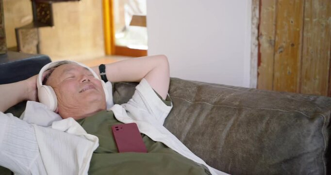 Pullback showing senior male lying on sofa, donning headset and listening to red phone, relaxing