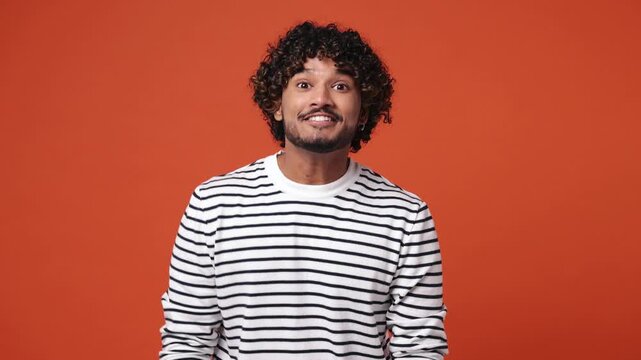 Young Indian man wear striped sweatshirt casual clothes look around for friend find wave meet greet with hand as notices someone say hello hi isolated on plain orange red background. Lifestyle concept