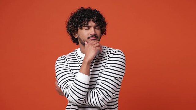 Puzzled young Indian man he wear casual clothes look aside put hand prop up on chin iterates over solution options feels doubtful thinks but no decision comes isolated on plain orange red background