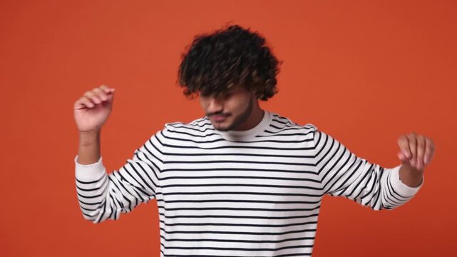 Young Indian man wear striped sweatshirt casual clothes dance wave expressive gesticulating hands fool around have fun enjoy isolated on plain orange red background studio portrait. Lifestyle concept