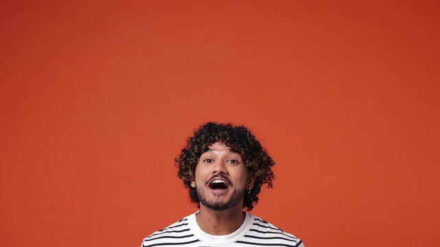 Young Indian man wear striped sweatshirt casual clothes pointing indicate index finger overhead on workspace area copy space isolated on plain orange red background studio portrait. Lifestyle concept